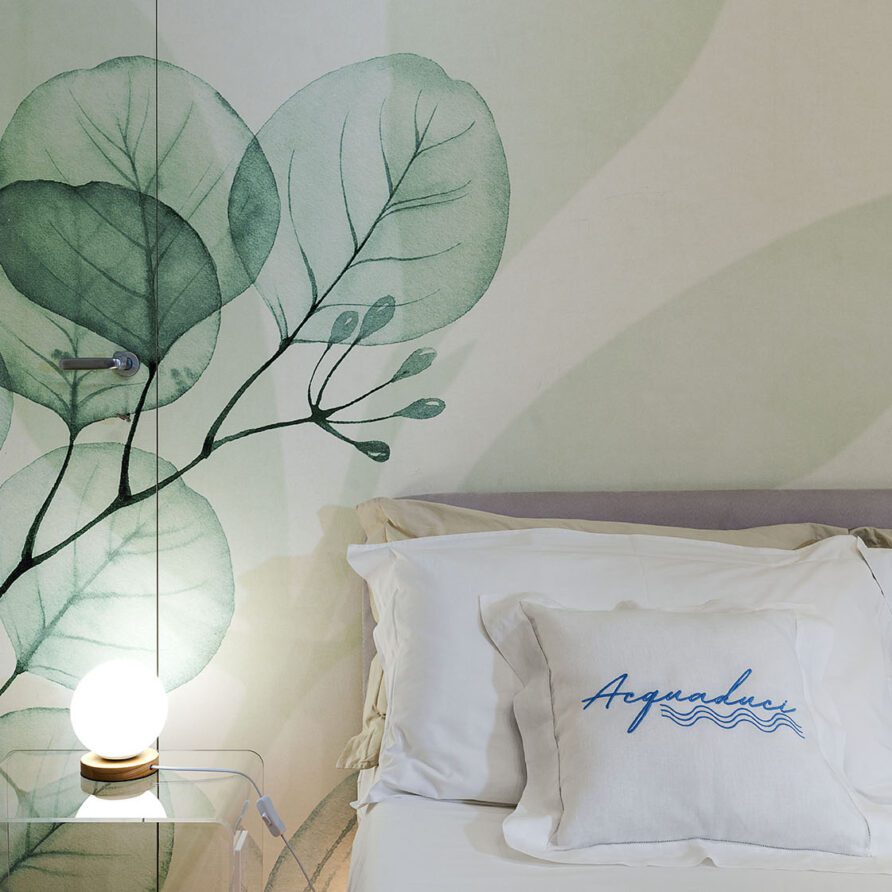 Dettaglio di un angolo di stanza con parete decorata con foglie verdi e letto con cuscino con scritta Artesa
