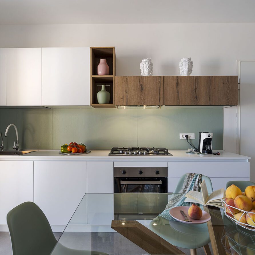 Cucina moderna con mobili bianchi, scaffale in legno, piano cottura e frigorifero in un ambiente luminoso