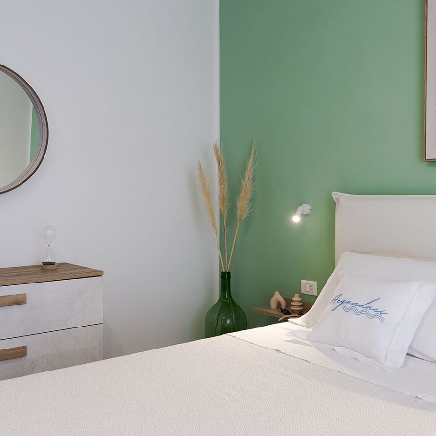Stanza moderna con pareti verdi, letto matrimoniale con cuscino decorativo e specchio rotondo sopra un comò bianco