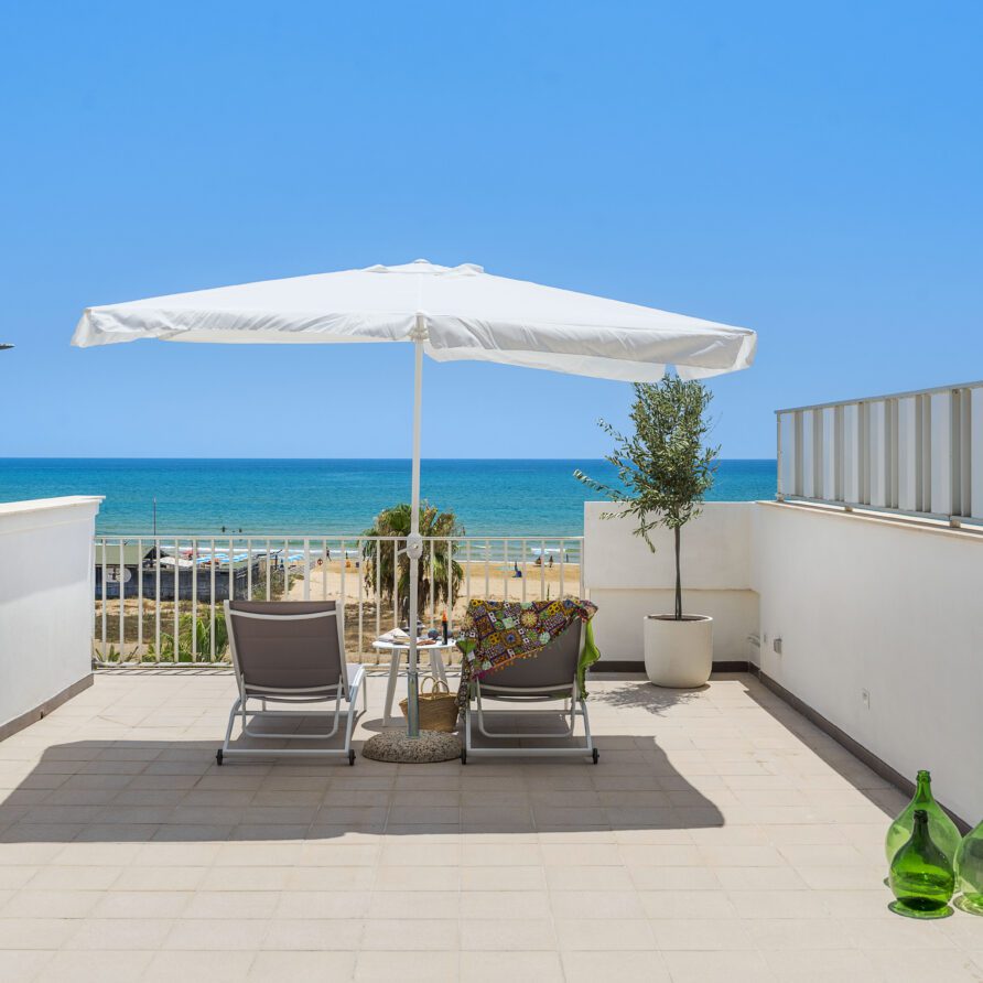 Terrazza in stile mediterraneo con sedie a sdraio, ombrellone e vista sul mare blu e spiaggia di sabbia