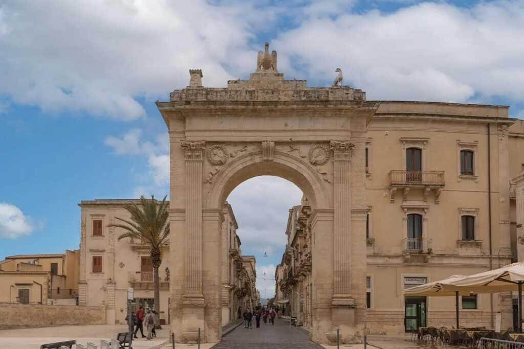 Arco di accesso storico con edifici antichi e palme sotto un cielo nuvoloso a Cagliari