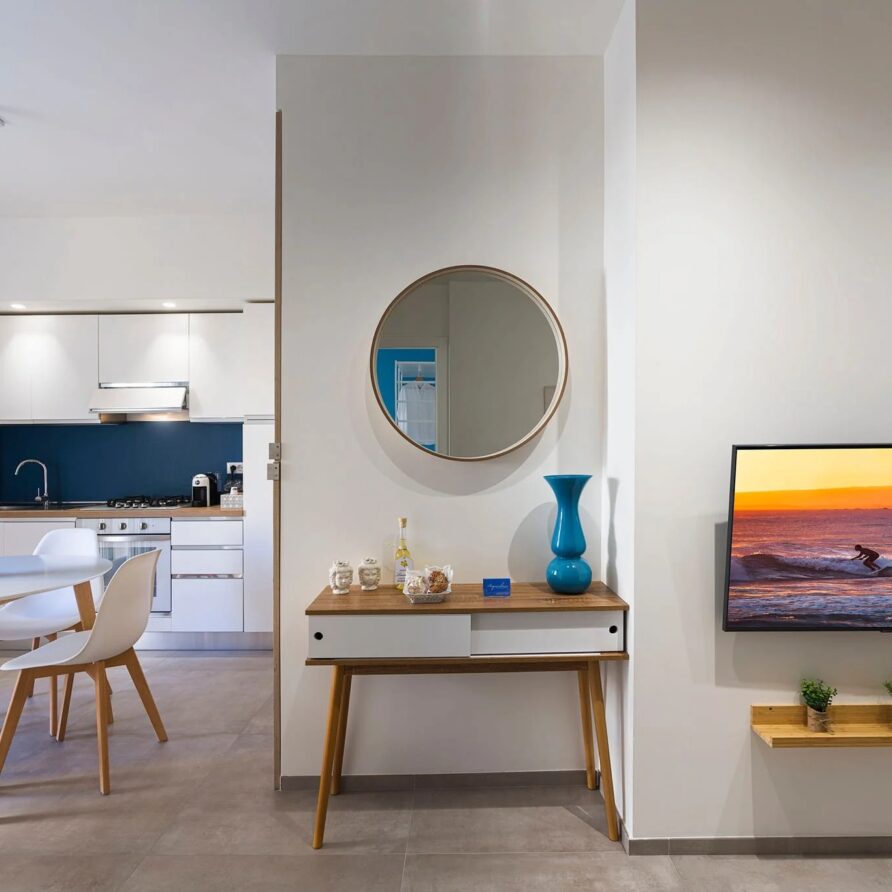 Interno di un soggiorno con parete bianca, tavolo da pranzo con sedie, specchio rotondo, consolle con decorazioni e un televisore con vista al tramonto