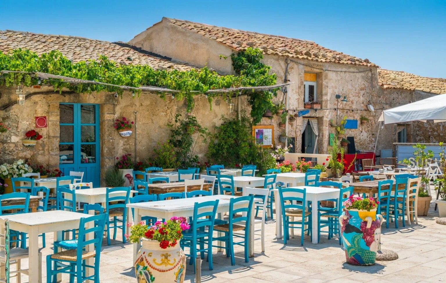 Ristorante all'aperto con tavoli e sedie azzurri in una pittoresca località mediterranea con edificio storico e decorazioni floreali