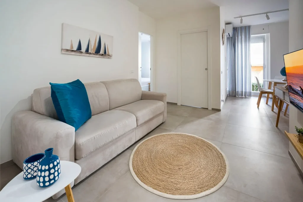 imgi_49_p Interno di soggiorno luminoso con divano beige, cuscini blu, tappeto rotondo in juta, e area relax con TV e balcone