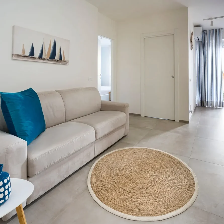 Interno di soggiorno luminoso con divano beige, cuscini blu, tappeto rotondo in juta, e area relax con TV e balcone