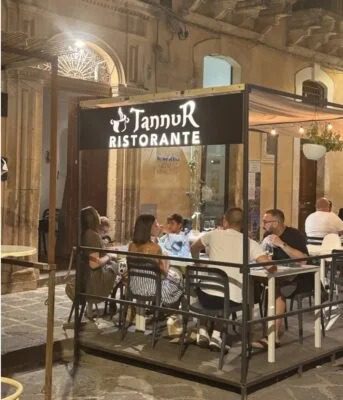 Gruppo di persone che mangiano all'aperto in un ristorante con insegna Tannur Ristorante all'ingresso