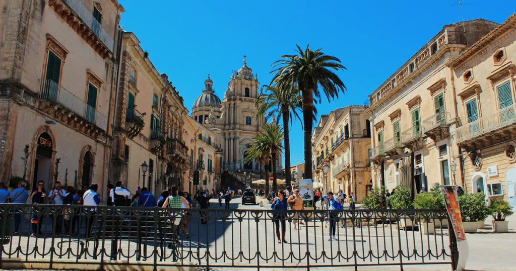 Vista di una piazza storica a Malta con palme, edifici antichi e un'abbazia imponente sullo sfondo
