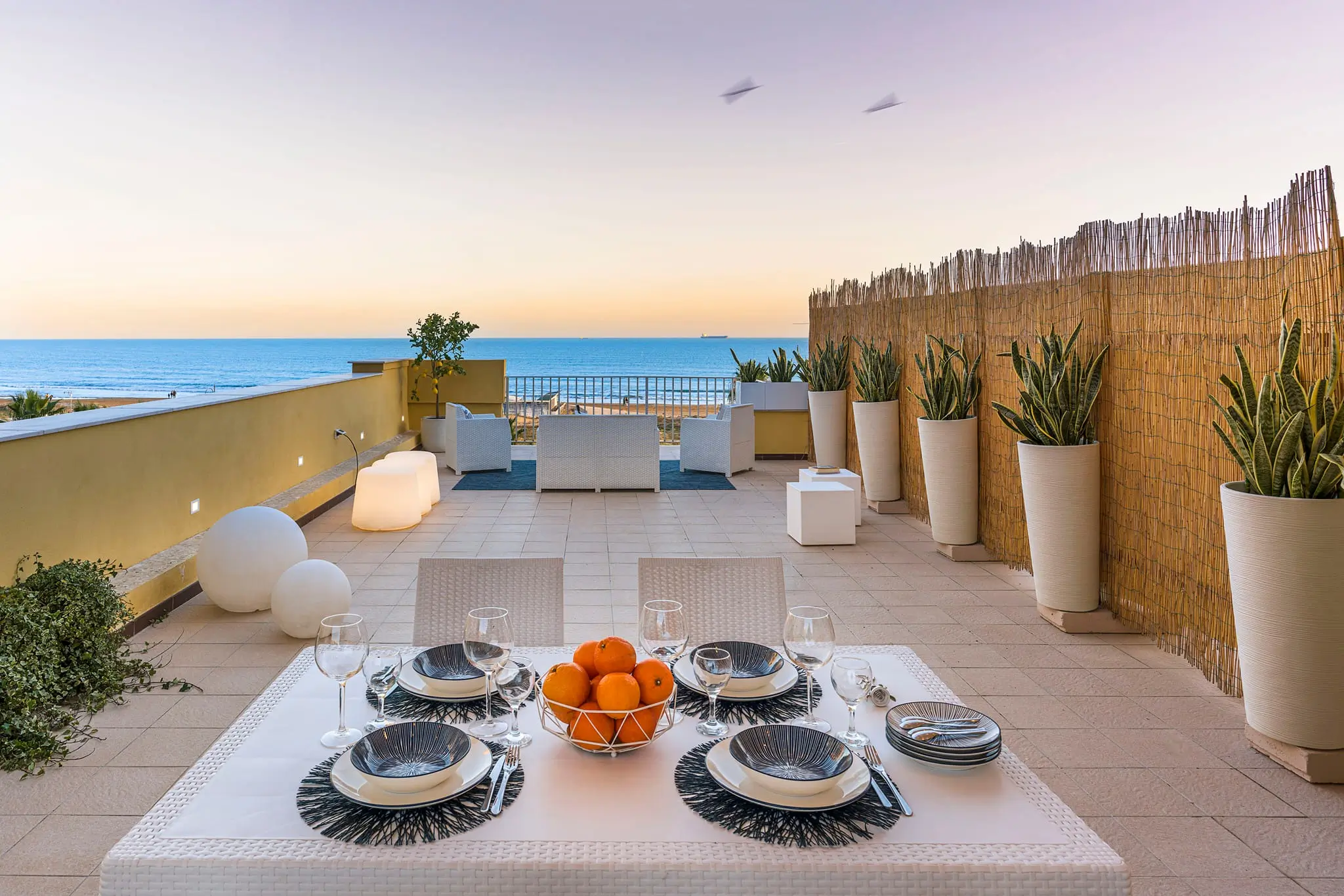Terrazza elegante con vista mare, tavola imbandita con frutta e stoviglie, piante in vasi e sedie comode per relax outdoor